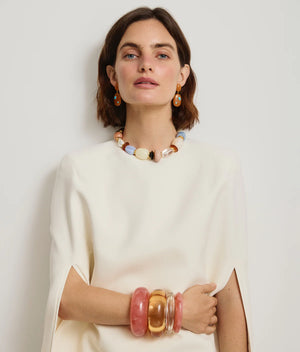 A woman in a cream top poses against a plain background, accessorized with chunky colorful jewelry including statement earrings, a necklace, and the Lizzie Fortunato Arc Marbled Cuff in pink resin.