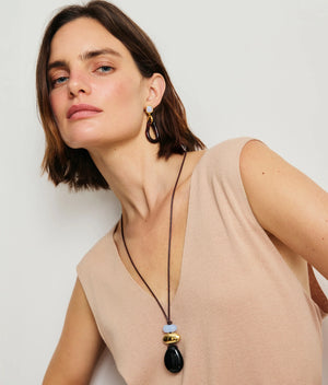 Woman with short brown hair models the Lizzie Fortunato Galina Earrings—gold and black statement pieces—paired with a sleeveless beige top and long beaded necklace, set against a plain light background.