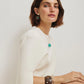 A woman with short brown hair wears a white dress, green topaz pendant necklace, Lizzie Fortunato Taj Disc Earrings, a brown bracelet, and a clear bangle, seated against a plain light background.