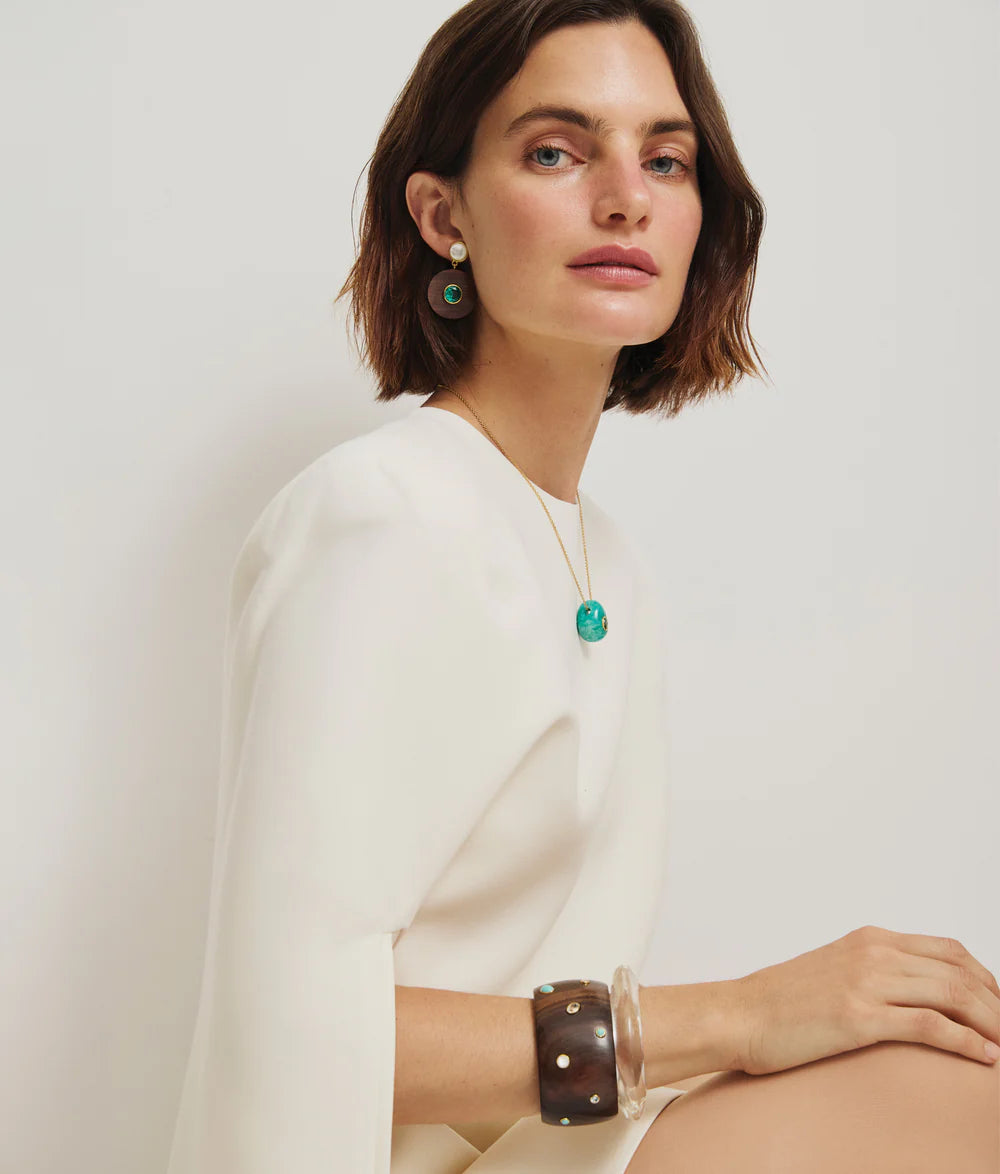 A woman with short brown hair wears a white dress, green topaz pendant necklace, Lizzie Fortunato Taj Disc Earrings, a brown bracelet, and a clear bangle, seated against a plain light background.