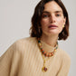 A woman with short brown hair wears a beige ribbed top, the Lizzie Fortunato Toro Earrings, and layered necklaces with beads and a gold pendant as she poses against a plain background.