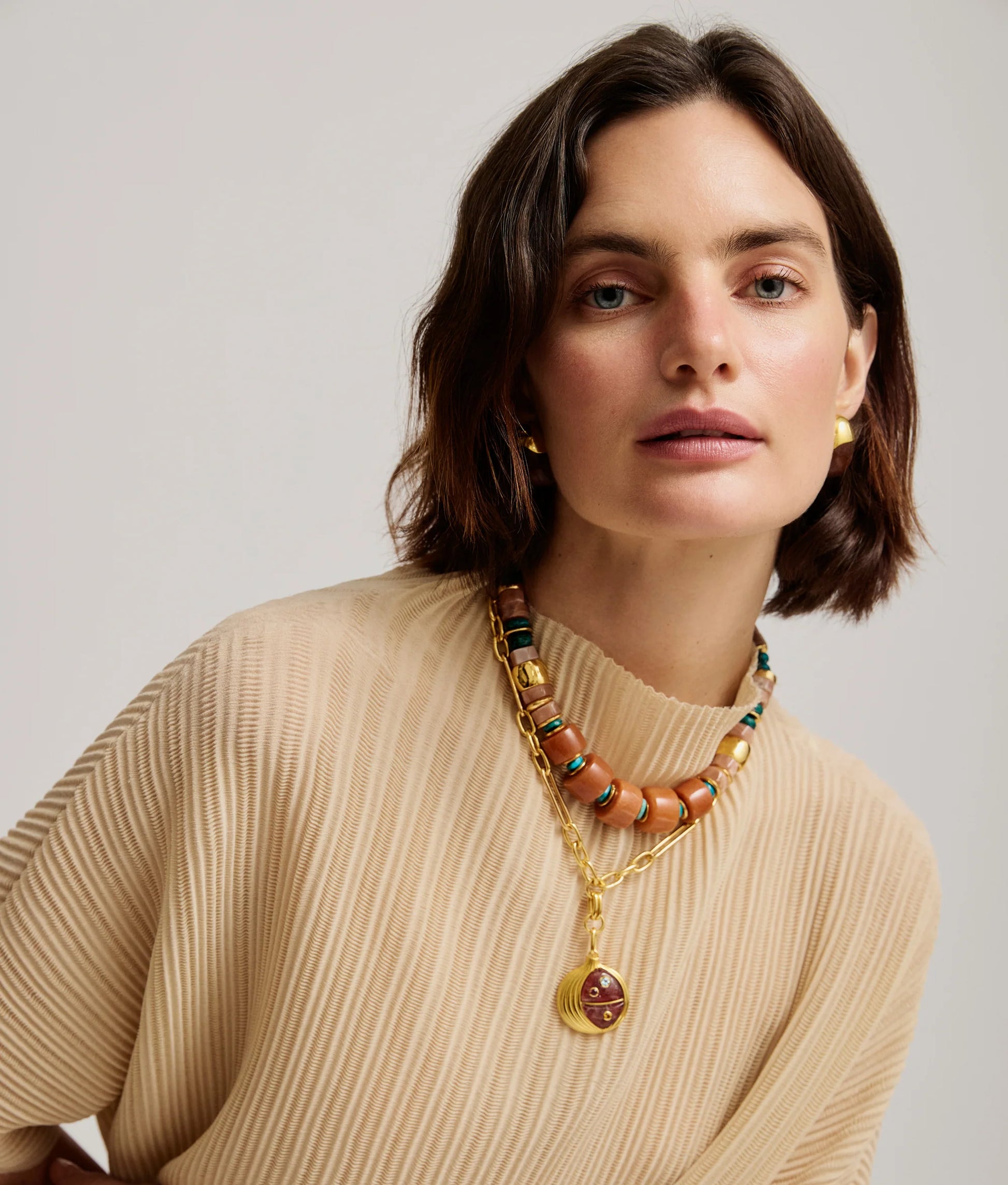 A woman with short brown hair wears a beige ribbed top, the Lizzie Fortunato Toro Earrings, and layered necklaces with beads and a gold pendant as she poses against a plain background.