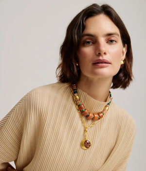 A woman with short brown hair wears a beige ribbed top, the Lizzie Fortunato Toro Earrings, and layered necklaces with beads and a gold pendant as she poses against a plain background.