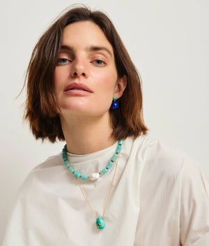 Woman with short brown hair wearing a white top and Lizzie Fortunato's Umbria Earrings, plus a turquoise beaded necklace and gold-plated brass pendant, against a plain light background.