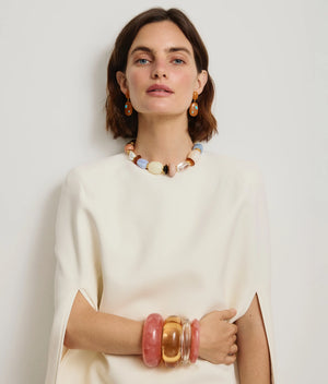 A woman in a white dress poses against a plain background, accessorized with chunky colorful jewelry including Lizzie Fortunato’s Satsuma Earrings, orange resin bracelets, and turquoise-colored stones.
