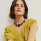 A woman with short brown hair poses against a plain light background wearing a yellow draped top, chunky beaded necklace, and Lizzie Fortunato's Laurel Earrings.