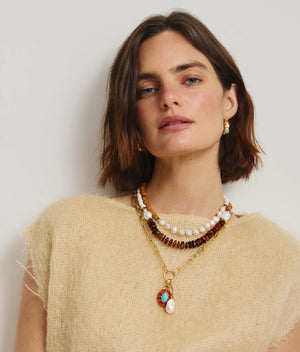 A woman with short brown hair wears Lizzie Fortunato's Piet Hoop Earrings, a beige textured top, and layered pearl, bead, and turquoise pendant necklaces, posing against a plain light background.
