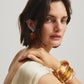 A woman with short dark hair wears a cream sleeveless top, large amber bangles, and Lizzie Fortunato Dulcinea Earrings—gold-plated brass adorned with turquoise—as she looks over her shoulder.
