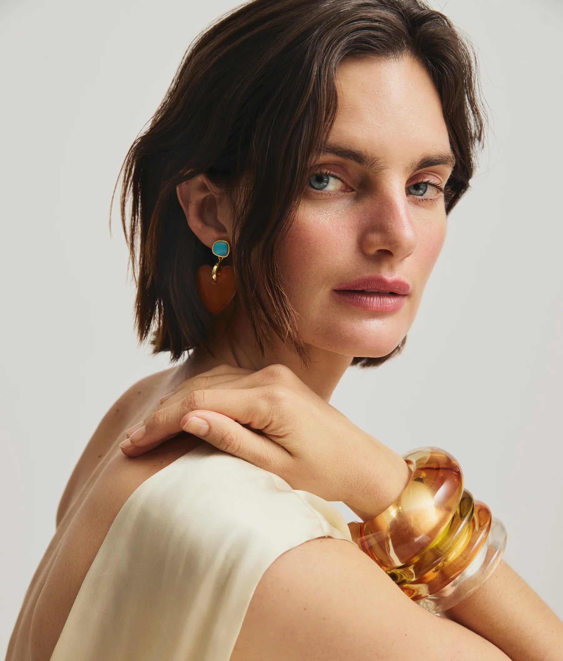 A woman with short dark hair wears a cream sleeveless top, large amber bangles, and Lizzie Fortunato Dulcinea Earrings—gold-plated brass adorned with turquoise—as she looks over her shoulder.