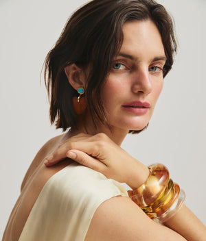 A woman with short dark hair wears a cream sleeveless top, large amber bangles, and Lizzie Fortunato Dulcinea Earrings—gold-plated brass adorned with turquoise—as she looks over her shoulder.