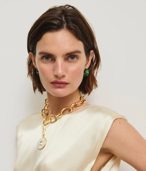 A woman with short brown hair wears a cream sleeveless top, a gold-plated brass chain necklace with a round pendant, and Lizzie Fortunato Zellige Hoops, against a plain light background.
