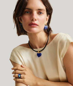 A woman with short brown hair wears a cream blouse and Lizzie Fortunato Dali Heart Earrings, along with matching blue heart-shaped glass jewelry, posing against a plain light background.