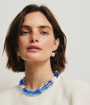 A woman with short brown hair wears a white top, a chunky blue and white beaded necklace, and one Lizzie Fortunato Pearl Pablo Earring (gold-plated sterling silver), looking at the camera against a plain background.