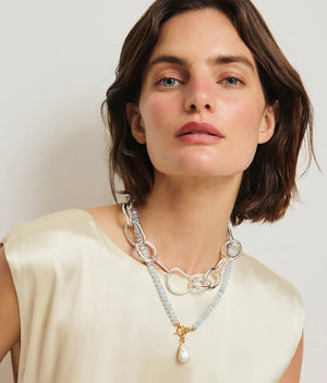 A woman with short brown hair in a cream sleeveless top and layered necklaces—including Lizzie Fortunato’s Pearl Isle Necklace—stands against a plain background.