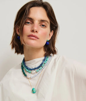 Woman with short brown hair wears the Lizzie Fortunato Cabana Necklace, a layered blue and green beaded piece, paired with a gold pendant necklace, blue earrings, and a white top, facing forward against a plain background.
