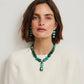 A woman with short brown hair wears a white top, turquoise stone drop earrings, and the Lizzie Fortunato Andamento Necklace with a bold turquoise pendant, posed against a plain light background.