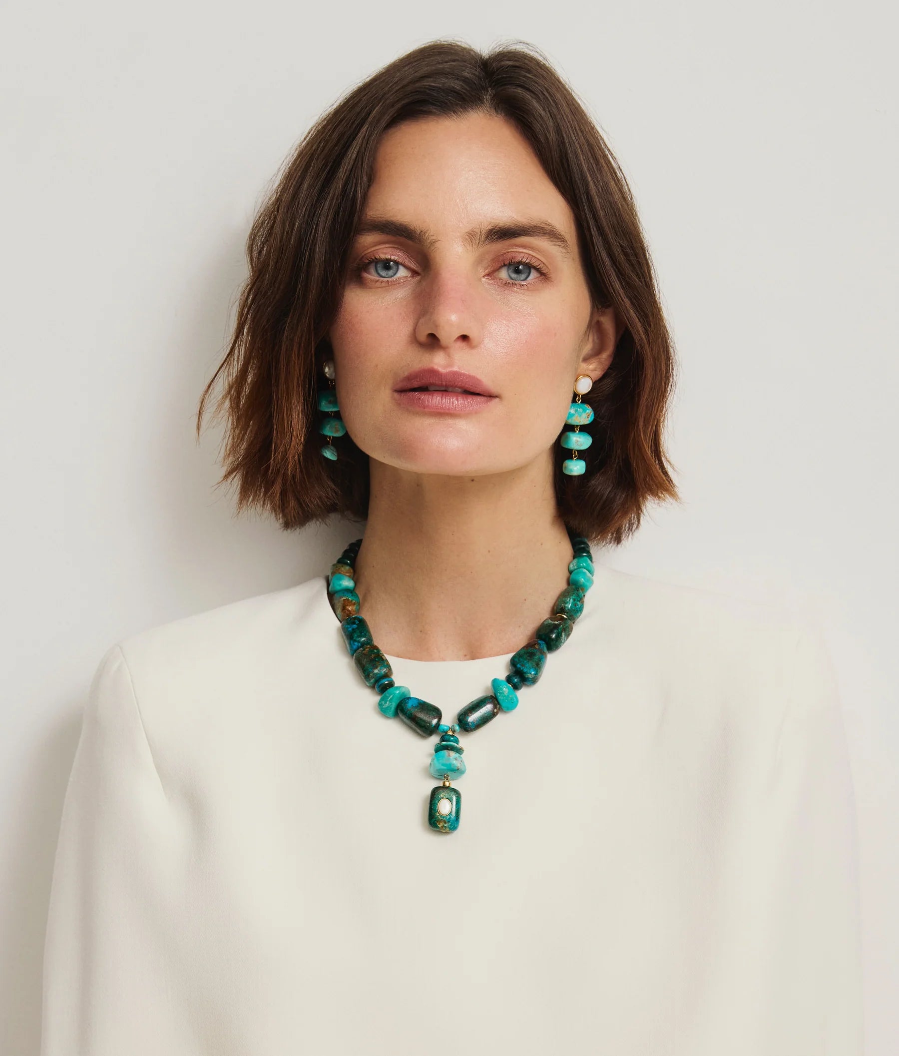 A woman with short brown hair wears a white top, turquoise stone drop earrings, and the Lizzie Fortunato Andamento Necklace with a bold turquoise pendant, posed against a plain light background.