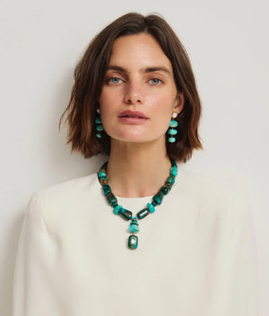 A woman with short brown hair wears a white top, turquoise stone drop earrings, and the Lizzie Fortunato Andamento Necklace with a bold turquoise pendant, posed against a plain light background.