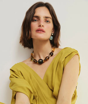A woman with short brown hair wears a yellow wrap dress and the Lizzie Fortunato Desert Mirage Collar necklace, along with a green earring, posing against a plain light-colored background.