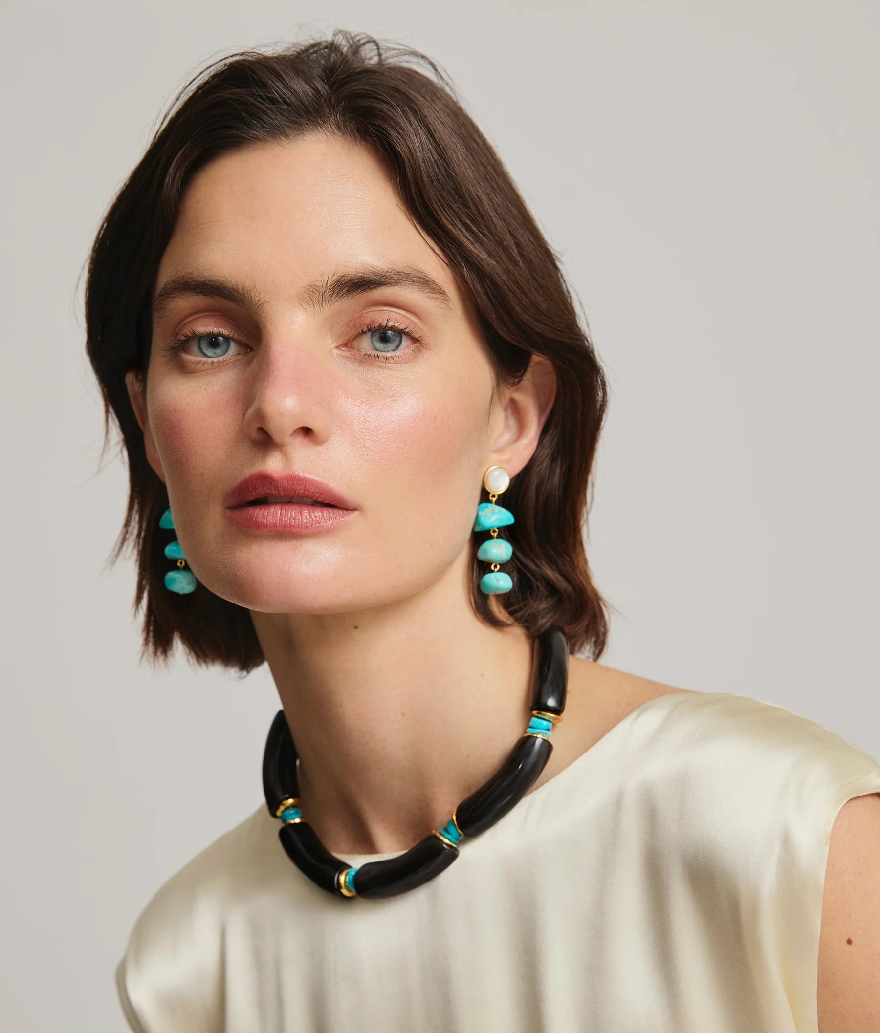 Woman with short brown hair wears a cream top and Lizzie Fortunato's Old World Earrings—gold-plated brass—plus a chunky black necklace with turquoise accents, looking at the camera against a plain background.