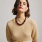 A woman stands against a neutral background wearing a beige, pleated dress with a belt, gold earrings, and the Lizzie Fortunato Amaranth Necklace—a single-strand amber heishi necklace with gold-plated beads.