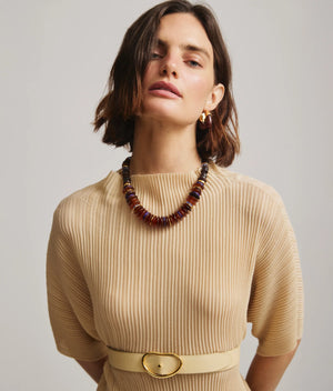 A woman stands against a neutral background wearing a beige, pleated dress with a belt, gold earrings, and the Lizzie Fortunato Amaranth Necklace—a single-strand amber heishi necklace with gold-plated beads.