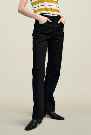 A person stands with hands in pockets against a plain background, wearing a yellow and white striped sleeveless top, the Kallmeyer Farah Classic Bootcut black stretch cotton jeans, and black pointed-toe shoes.