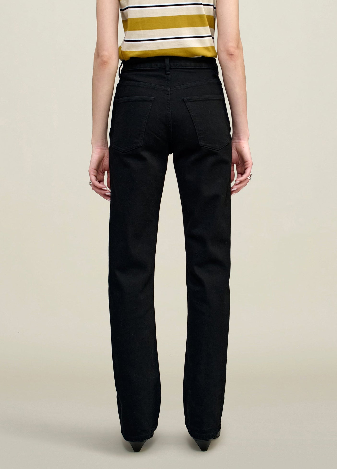 A person stands facing away, wearing a sleeveless striped top and the Kallmeyer Farah Classic Bootcut high-waisted black jeans with back pockets, crafted from stretch cotton denim, against a plain light background.