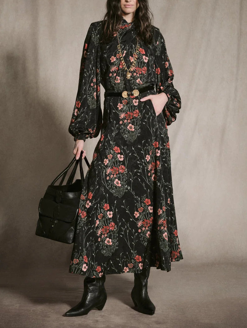 A person wearing a Johanna Ortiz Jacquard Encanto De Isla Ankle Skirt as a high-neck midi dress with a botanical motif, styled with a black belt, gold necklace, black boots, and a black tote bag.