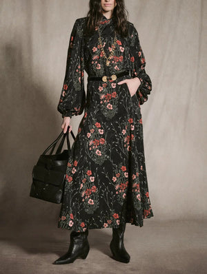A person wearing a Johanna Ortiz Jacquard Encanto De Isla Ankle Skirt as a high-neck midi dress with a botanical motif, styled with a black belt, gold necklace, black boots, and a black tote bag.