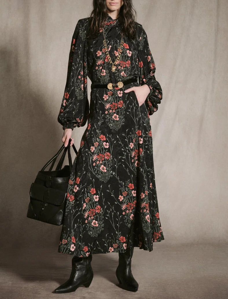 A person wearing a floor-length black Jacquard Intricate Islands Top with a botanical motif, a black belt with an ornate gold buckle, and black boots, holding a black leather tote bag.