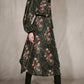 A woman wears a Johanna Ortiz Golden Vibe Dress in a long-sleeved, botanical print midi style, accessorized with a thin black belt and black cowboy boots.