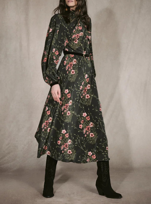 A woman wears a Johanna Ortiz Golden Vibe Dress in a long-sleeved, botanical print midi style, accessorized with a thin black belt and black cowboy boots.
