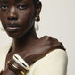 A woman wearing a cream top, pearl drop earrings, and two chunky bracelets—including the Halo Cuff by Lizzie Fortunato—poses with her hand on her shoulder against a neutral background.