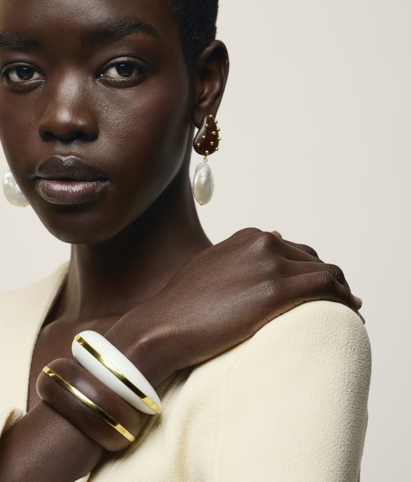 A woman wearing a cream top, pearl drop earrings, and two chunky bracelets—including the Halo Cuff by Lizzie Fortunato—poses with her hand on her shoulder against a neutral background.