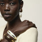 A woman wearing a cream top, pearl drop earrings, and two Lizzie Fortunato Halo Cuffs—one in white with gold trim and the other in brown with gold trim reminiscent of elegant acacia wood—poses against a neutral background.