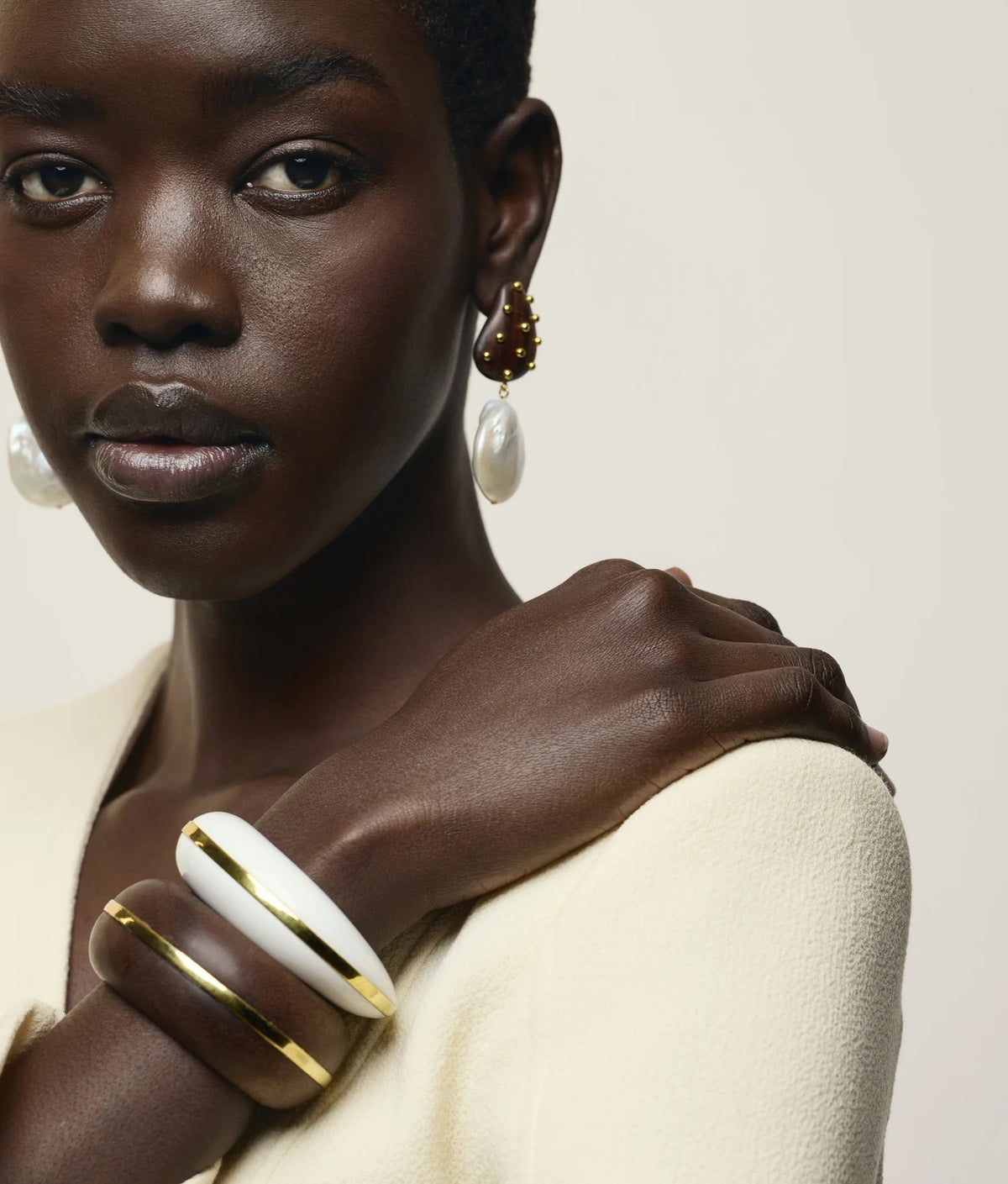 A woman wearing a cream top, pearl drop earrings, and two Lizzie Fortunato Halo Cuffs—one in white with gold trim and the other in brown with gold trim reminiscent of elegant acacia wood—poses against a neutral background.