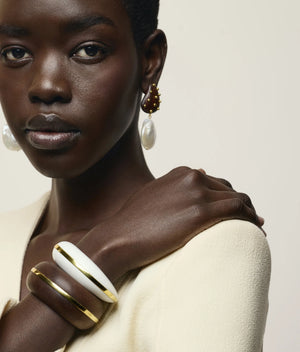 A woman wearing a cream top, pearl drop earrings, and two Lizzie Fortunato Halo Cuffs—one in white with gold trim and the other in brown with gold trim reminiscent of elegant acacia wood—poses against a neutral background.