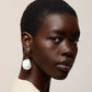A person with short hair wears a cream top and the Lizzie Fortunato Orbit Earrings, featuring large freshwater pearl drops, looking over their shoulder against a plain, neutral background.
