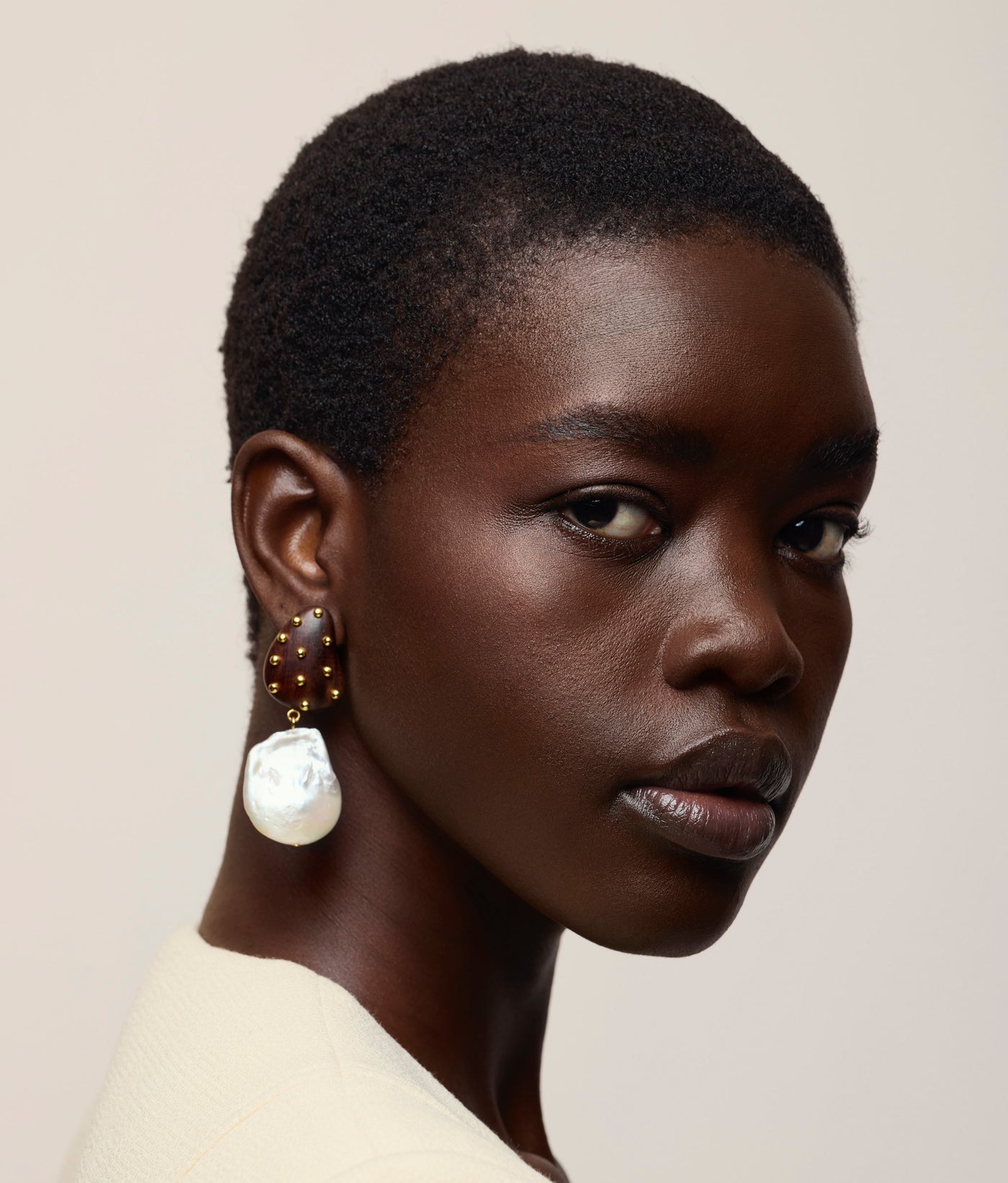 A person with short hair wears a cream top and the Lizzie Fortunato Orbit Earrings, featuring large freshwater pearl drops, looking over their shoulder against a plain, neutral background.
