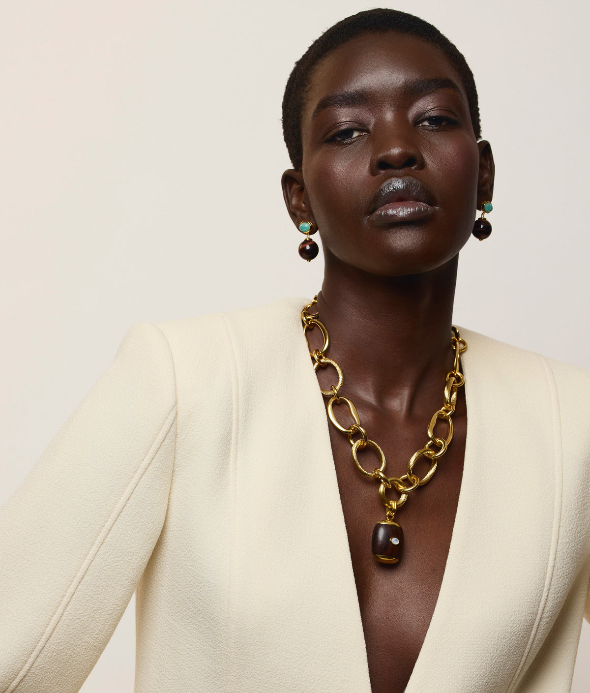 A person with short hair wears a cream blazer, a chunky gold necklace with a dark pendant, and striking Lizzie Fortunato Umbria Earrings that perfectly complement the look.