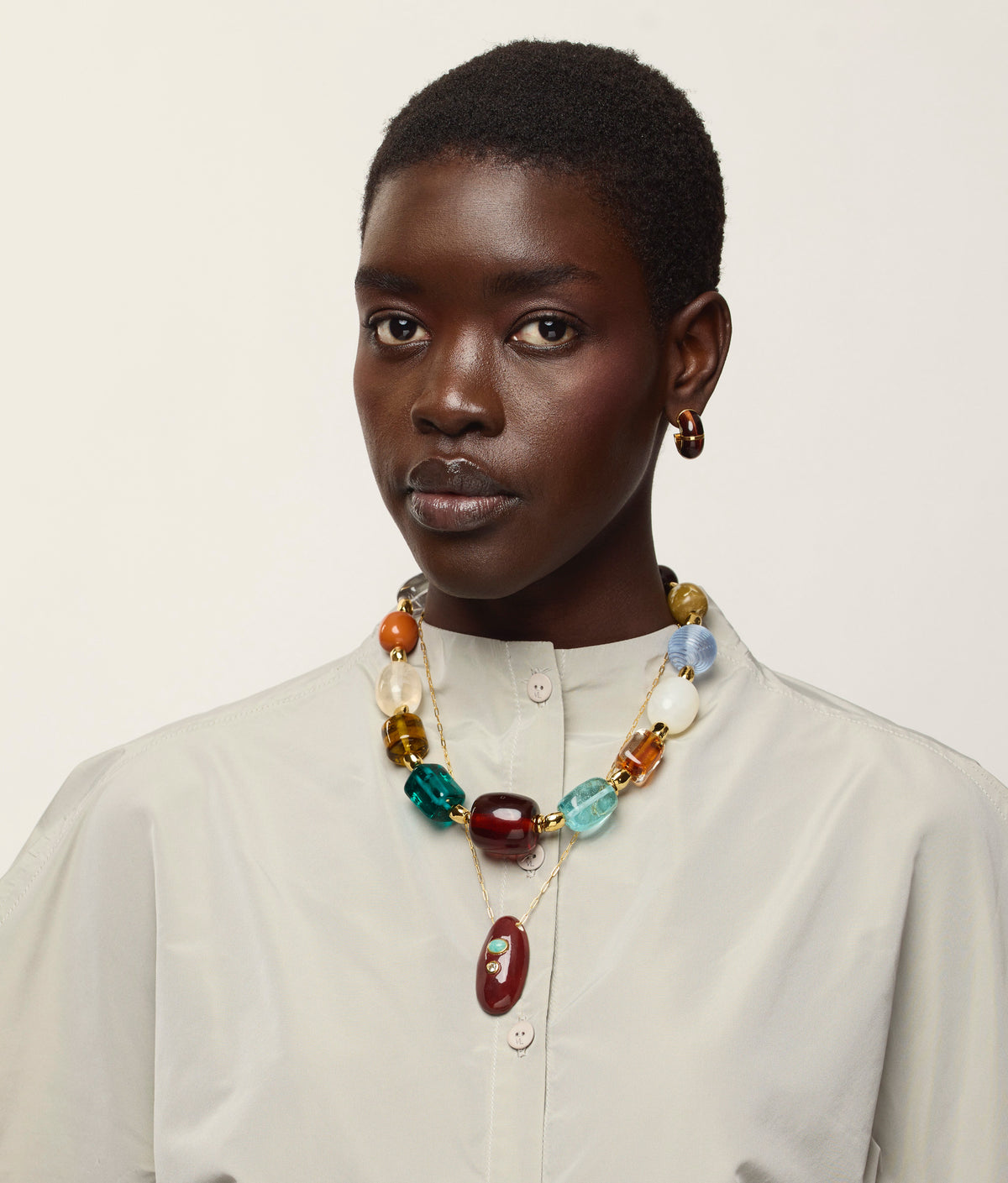A person wearing a cream button-up shirt, Lizzie Fortunato Zellige Hoops, and a colorful chunky beaded necklace stands against a plain light background.