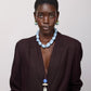 A person wearing a dark blazer and the Lizzie Fortunato Sea and Sky Collar—a large statement necklace featuring light blue amazonite beads—poses against a plain light background, styled with drop earrings and a long pendant necklace.