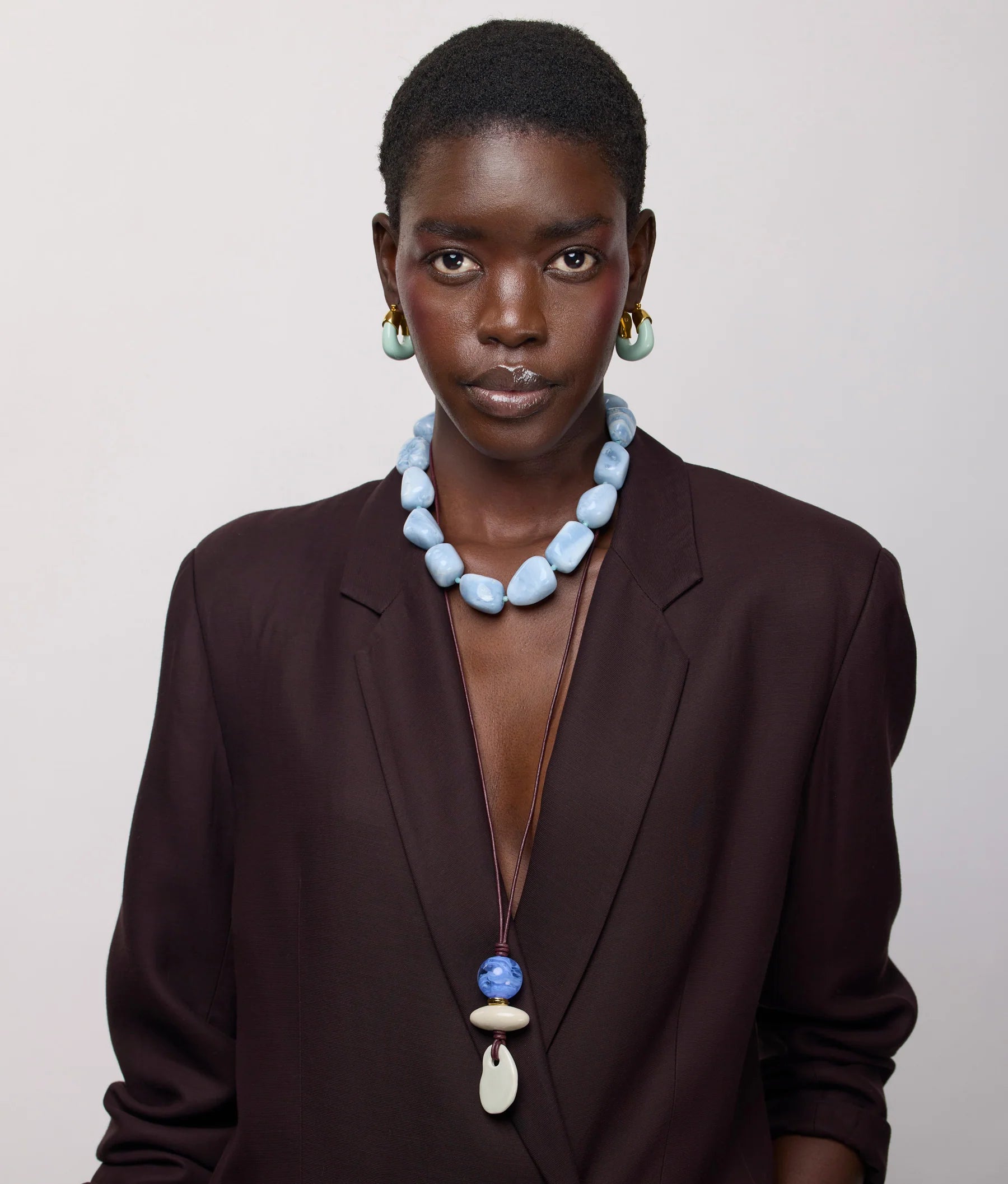 A person wearing a dark blazer and the Lizzie Fortunato Sea and Sky Collar—a large statement necklace featuring light blue amazonite beads—poses against a plain light background, styled with drop earrings and a long pendant necklace.