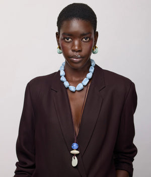 A person wearing a dark blazer and the Lizzie Fortunato Sea and Sky Collar—a large statement necklace featuring light blue amazonite beads—poses against a plain light background, styled with drop earrings and a long pendant necklace.