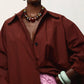 A person wearing a loose maroon coat, the Lizzie Fortunato Carmine Collar—a statement necklace with gold-plated beads—large hoop earrings, green bangles, and a crinkled pink skirt. Only the lower face and upper body are visible.