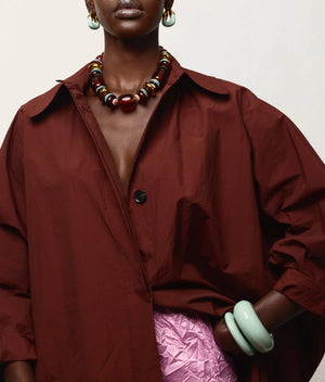 A person wearing a loose maroon coat, the Lizzie Fortunato Carmine Collar—a statement necklace with gold-plated beads—large hoop earrings, green bangles, and a crinkled pink skirt. Only the lower face and upper body are visible.
