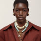 A person with short hair wears a maroon shirt, gold earrings, and the Lizzie Fortunato Desert Bloom Necklace—layered beaded strands featuring amazonite beads and a delicate flower charm with a gold pendant—set against a plain background.