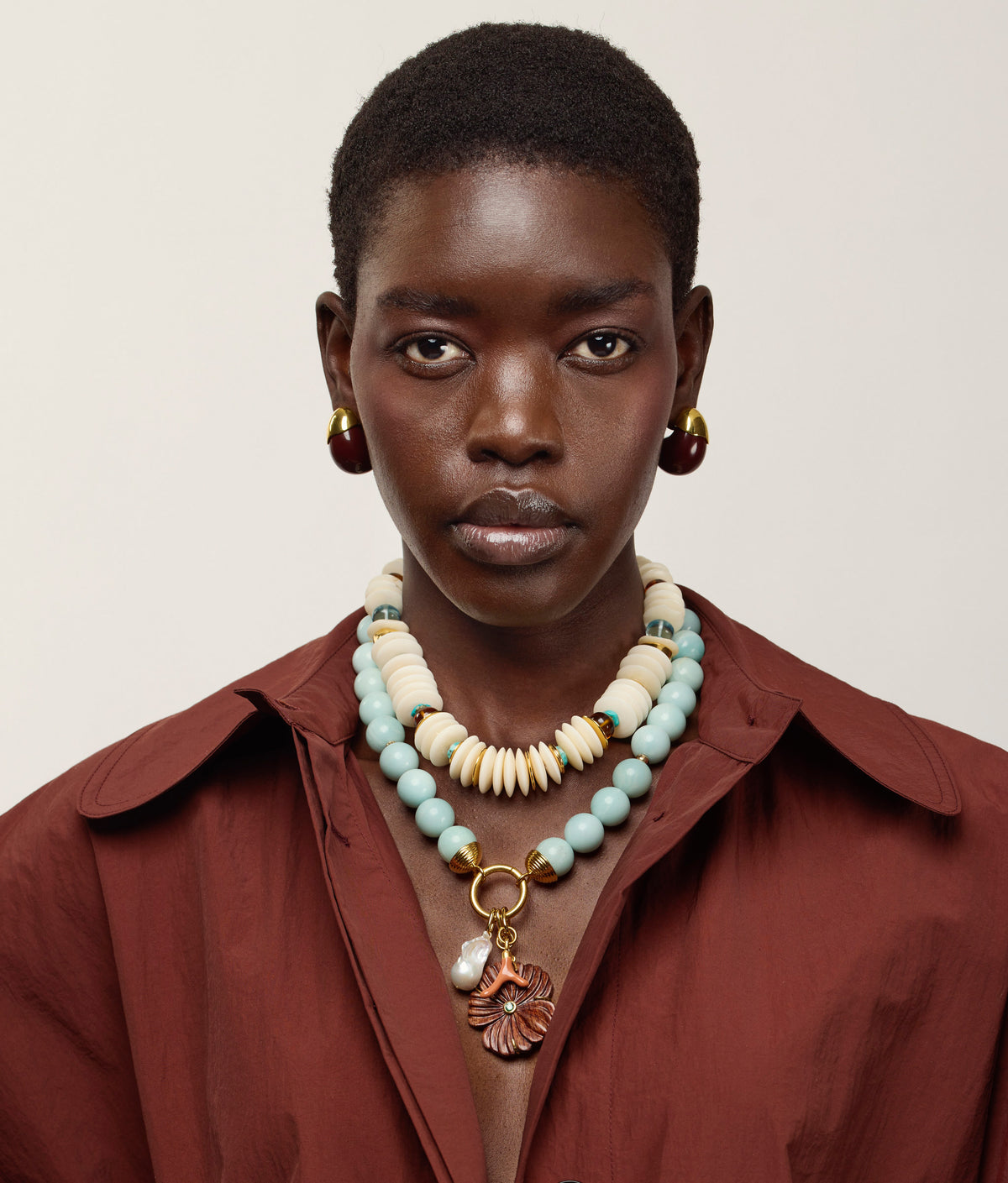 A person with short hair wears a maroon shirt, gold earrings, and the Lizzie Fortunato Desert Bloom Necklace—layered beaded strands featuring amazonite beads and a delicate flower charm with a gold pendant—set against a plain background.