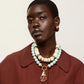 A woman with short hair wears a maroon coat and the Lizzie Fortunato Petra Collar—a large, layered necklace crafted from recycled white bone beads with gold-plated accents—along with a single oval earring, looking at the camera against a plain background.